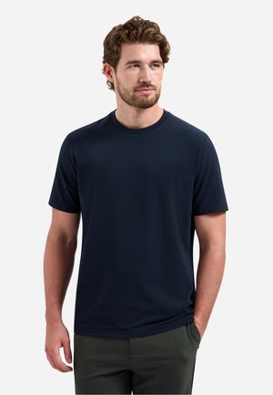 Man met bruin haar en baard, gekleed in een effen donkerblauw T-shirt met korte mouwen en donkere broek, staand met één hand in de zak.