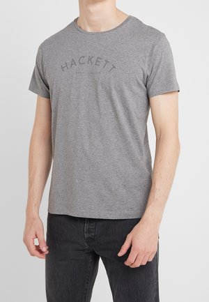 Mann in einem grauen Kurzarm-T-Shirt mit Hackett London England-Logo und einer schwarzen Jeans, der vor einem schlichten weißen Hintergrund steht.