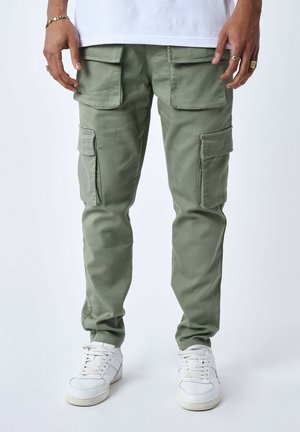 Pantalons cargo en tissu vert clair avec plusieurs poches latérales. Coupe slim avec des poignets élastiqués, portés avec des baskets blanches.