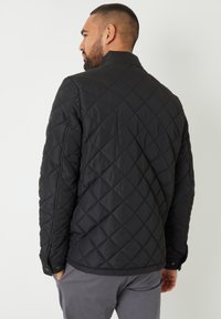 Homme avec une barbe courte portant une veste matelassée noire et un pantalon gris, se tenant de dos avec les mains dans les poches.