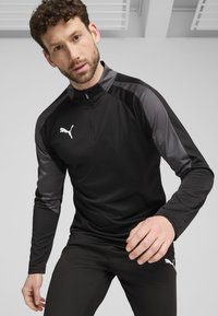 Svart atletisk zip-up topp med grå aksenter, lange ermer og høy krage. Har en hvit logo på brystet. Glatt, lett materiale.
