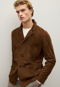 Giacca in suede marrone con colletto, due tasche frontali e chiusura con bottoni. Abbinata a un maglione bianco e uno sfondo testurizzato.