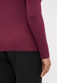 Long-sleeve maroon shirt with a smooth texture, featuring a fitted cut and a seamless hem, paired with black fabric pants.