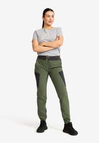 Woman stands with arms crossed wearing grey logo t-shirt, green cargo pants with black pockets, and black sneakers on plain white background.