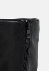 Sac en simili cuir noir avec une surface texturée et une fermeture éclair lisse, doté d'un dessus fin et d'un détail de fermeture éclair verticale sur un côté.