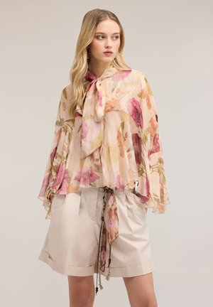 Jonge vrouw draagt een doorschijnende bloemenblouse met een gestrikte strik bij de hals en losse beige korte broek met een bloemenriem, poserend tegen een effen achtergrond.