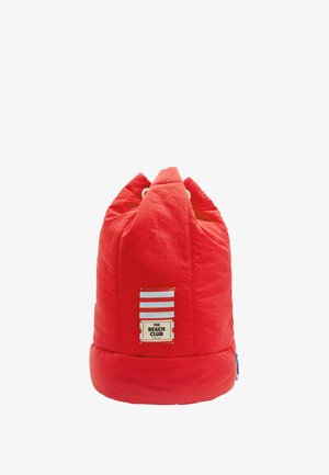 Czerwony, pikowany worek z fasolą z zaokrągloną podstawą, dwoma szczytami i naszywką z logo przedstawiającą niebieskie i białe paski. Teksturowana tkanina z widocznymi szwami.