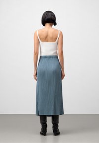 Vit åtsittande linne, sett bakifrån, kombinerat med en lång plisserad blå kjol. Kjolen har en subtil lyster och en hög midja, kompletterad med svarta stövlar.