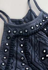 Haut sans manches bleu foncé avec boutons recouverts de tissu et motifs brodés complexes sur un fond clair.