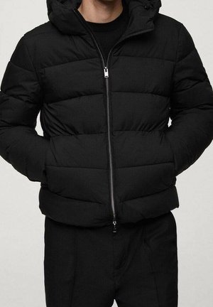 Mężczyzna ubrany w czarną pikowaną kurtkę puchową z przednim zamkiem i rękami w bocznych kieszeniach, w połączeniu z czarnymi spodniami.