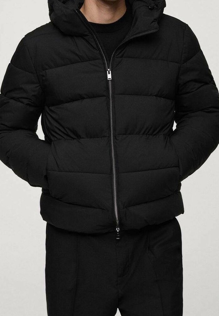 Homme portant une doudoune noire matelassée avec une fermeture éclair à l'avant et les mains dans les poches latérales, associée à un pantalon noir.
