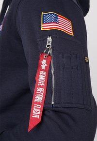 Hanorac bleu marin cu un patch cu steagul american, buzunar lateral cu fermoar și etichetă roșie din material cu textul "ÎNLEFRAȚI ÎNAINTE DE ZBOR".