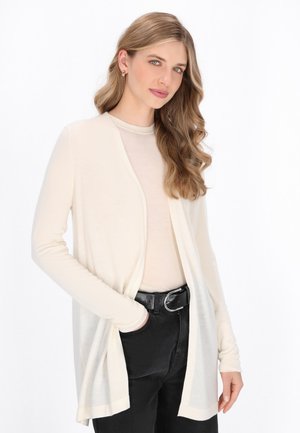 Lichte crème cardigan gemaakt van zachte materialen met een open voorkant, lange mouwen en een ontspannen pasvorm, gedragen over een beige top en zwarte broek.