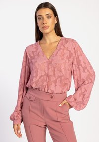 Rosa blumengemusterte Bluse mit V-Ausschnitt, langen Ärmeln und transparenter Stoffqualität. Das Model trägt maßgeschneiderte rosa Hose mit Knopfdetails.