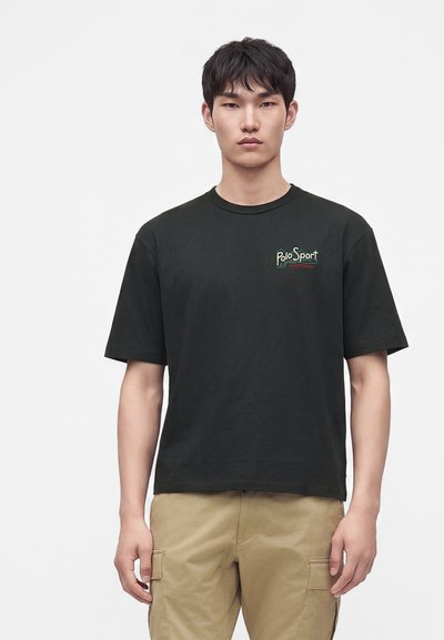 Νέος άνδρας με μαύρο T-shirt Polo Sport και μπεζ cargo παντελόνι, στέκεται σε απλό λευκό φόντο.