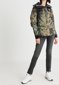 Chaqueta de camuflaje en verde y negro con capucha, con cierre de cremallera y botón, combinada con jeans grises y zapatillas blancas.