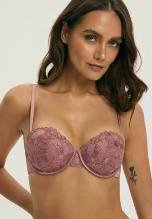 Kobieta z brązowymi włosami do ramion, nosząca różowo-fioletowy kwiecisty koronkowy stanik, na jej górnej części ramienia widoczny jest niewielki tatuaż czarnej róży.