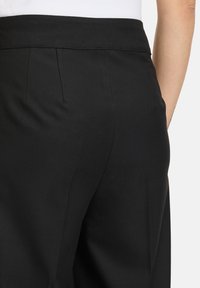 Pantalons noirs ajustés avec un tissu lisse, comportant une taille dissimulée et des poches latérales. Des plis ajoutent un détail structuré.