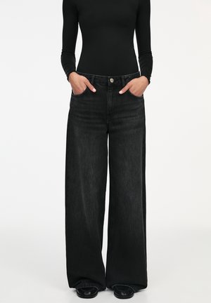 Personne portant un jean noir taille haute à jambes larges, les mains dans les poches, associé à un haut noir ajusté à manches longues et à des chaussures noires.