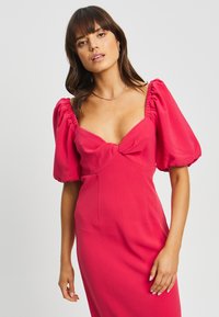 Abito fucsia aderente con maniche a sbuffo, scollatura a cuore e dettaglio nodo sul petto. Realizzato in tessuto morbido e leggero.