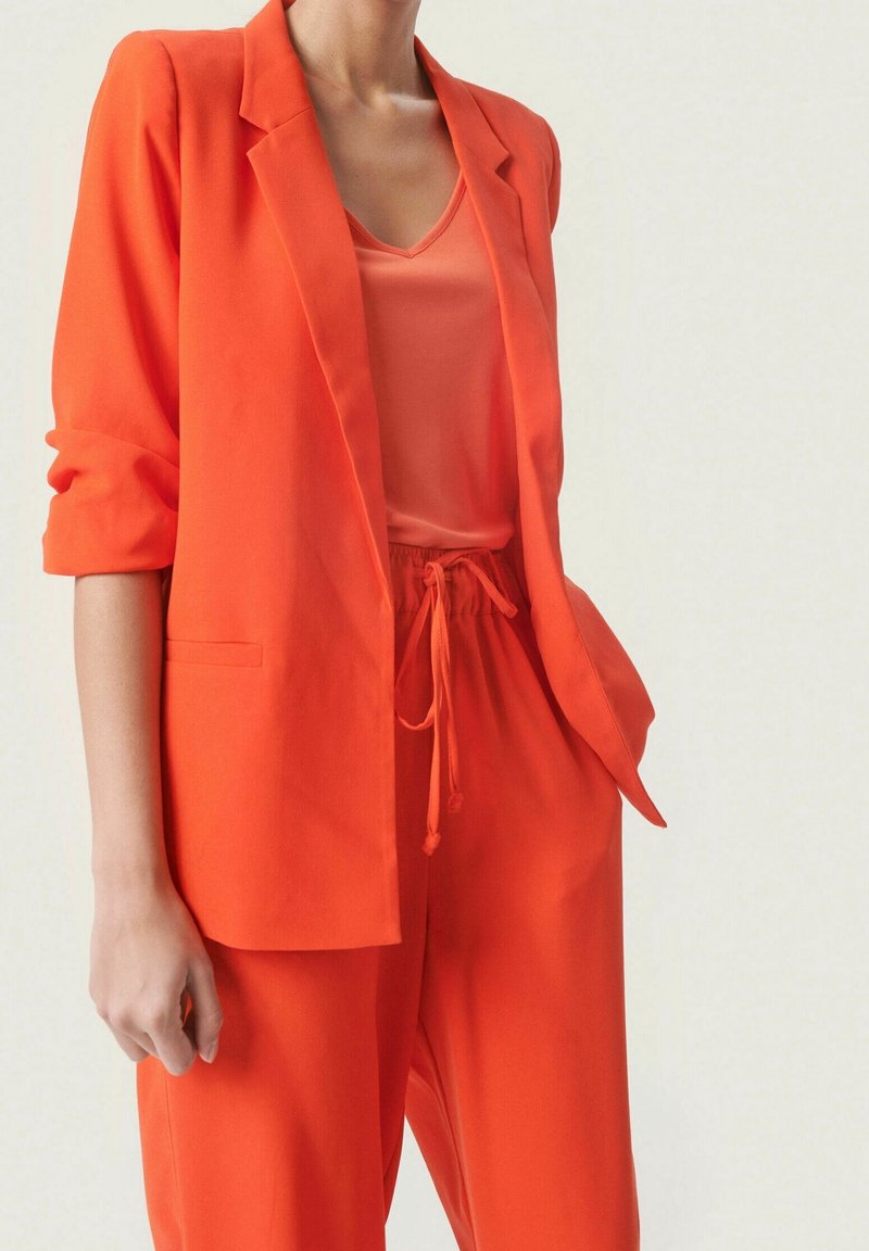 Pomarańczowa marynarka dopasowana z podwiniętymi rękawami, w zestawieniu z dopasowaną bluzką i spodniami na sznurku, podkreślająca spójną, monochromatyczną stylizację.