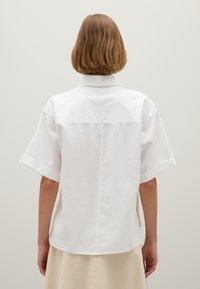White short-sleeve shirt with a relaxed fit, buttoned collar, and visible seam details. Behind view shows flat-laying hem and cuffed sleeves.