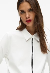 Balts garajmaka polo krekls ar melnu logotipa joslu priekšā, klasiskā apkakle un pogām. Mīksta kokvilnas materiāls.