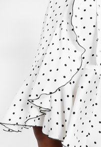 Vit kjol med svarta prickar, med en volangkant och kontrasterande svart sömnad längs kanten. Slät tygkvalitet.