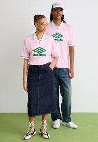 Duas modelos vestem camisolas cor-de-rosa com gola da Umbro, com logotipos verdes. Uma usa uma saia de ganga longa; a outra está com jeans de ganga casuais e ténis.