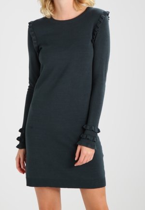 Długa czarna sukienka z długim rękawem, ozdobiona falbankami na ramionach i mankietach, noszona przez osobę na jasnym, jednolitym tle.