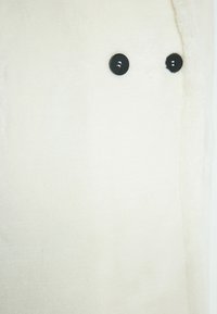 Krémová bunda z měkkého, plyšového materiálu; má jako ozdobu dvě černé knoflíky podél otevírání. Hladká, teplá textura.