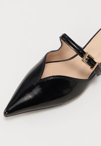 Chaussure en cuir verni noir à bout pointu avec un design découpé et une sangle à la cheville ornée d'une boucle dorée. Doublure intérieure lisse.