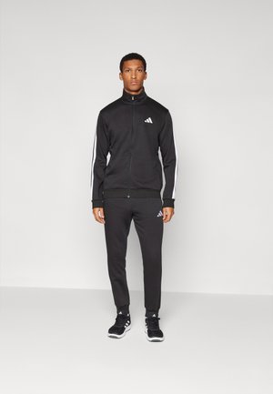 Chándal atlético negro que incluye una chaqueta con cremallera con franjas blancas y un logo, combinado con pantalones ajustados a juego y zapatillas negras.
