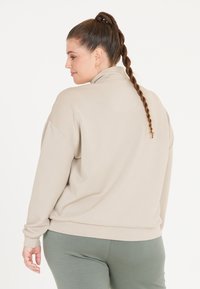 Beige höghalset sweatshirt med långa ärmar, mjukt material och avslappnad passform. Bärs med åtsittande gröna leggings. Minimalistisk design.