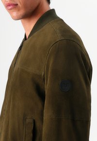 Λαδί  bomber jacket από απαλό σουέτ με ριμπ γιακά και μανίκια, που διαθέτει στρογγυλή μαύρη ετικέτα λογότυπου στον αριστερό μανσέτα.
