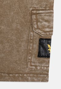 Bruine stof met een gestikt zakflapje en een zwart rechthoekig stukje met een geel vogel-embleem aan de rechteronderkant.