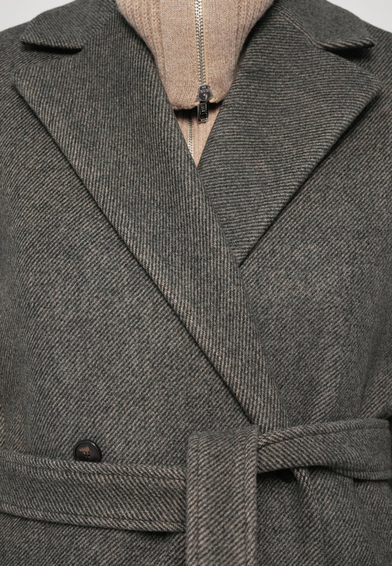 Grau gestreifter Mantel mit breiten Revers, einem Wickeldesign und einem passenden Bindegürtel. Darunter ist ein beiges Reißverschluss-Sweatshirt sichtbar.