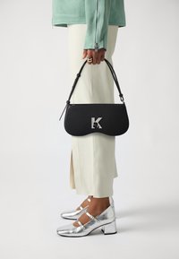 Borsa a spalla in pelle nera con forma curvata, hardware con la lettera 'K' in argento, abbinata a un top verde menta e pantaloni beige su misura. Tacchi block in argento.