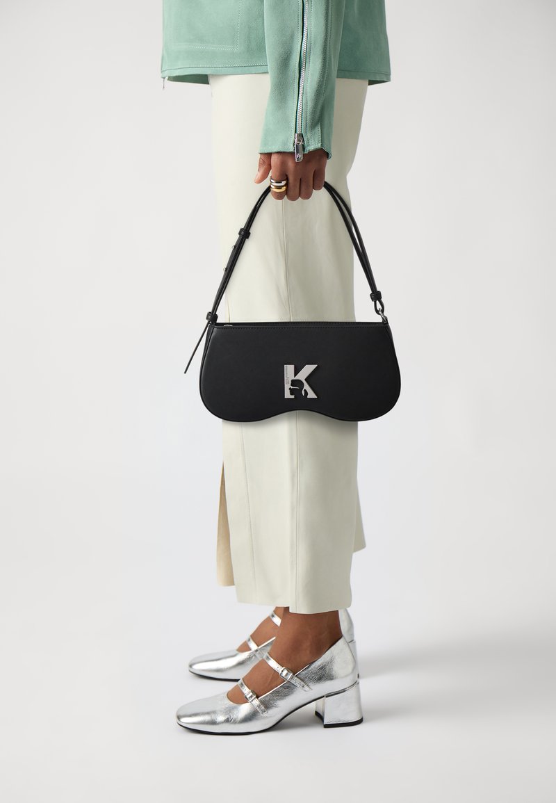 Borsa a spalla in pelle nera con forma curvata, hardware con la lettera 'K' in argento, abbinata a un top verde menta e pantaloni beige su misura. Tacchi block in argento.