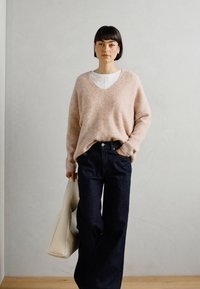 Hellrosafarbener, locker sitzender Strickpullover mit V-Ausschnitt, kombiniert mit dunkelblauen, weit geschnittenen Jeans. Das Model hält eine cremefarbene Tragetasche. Glatte Textur.