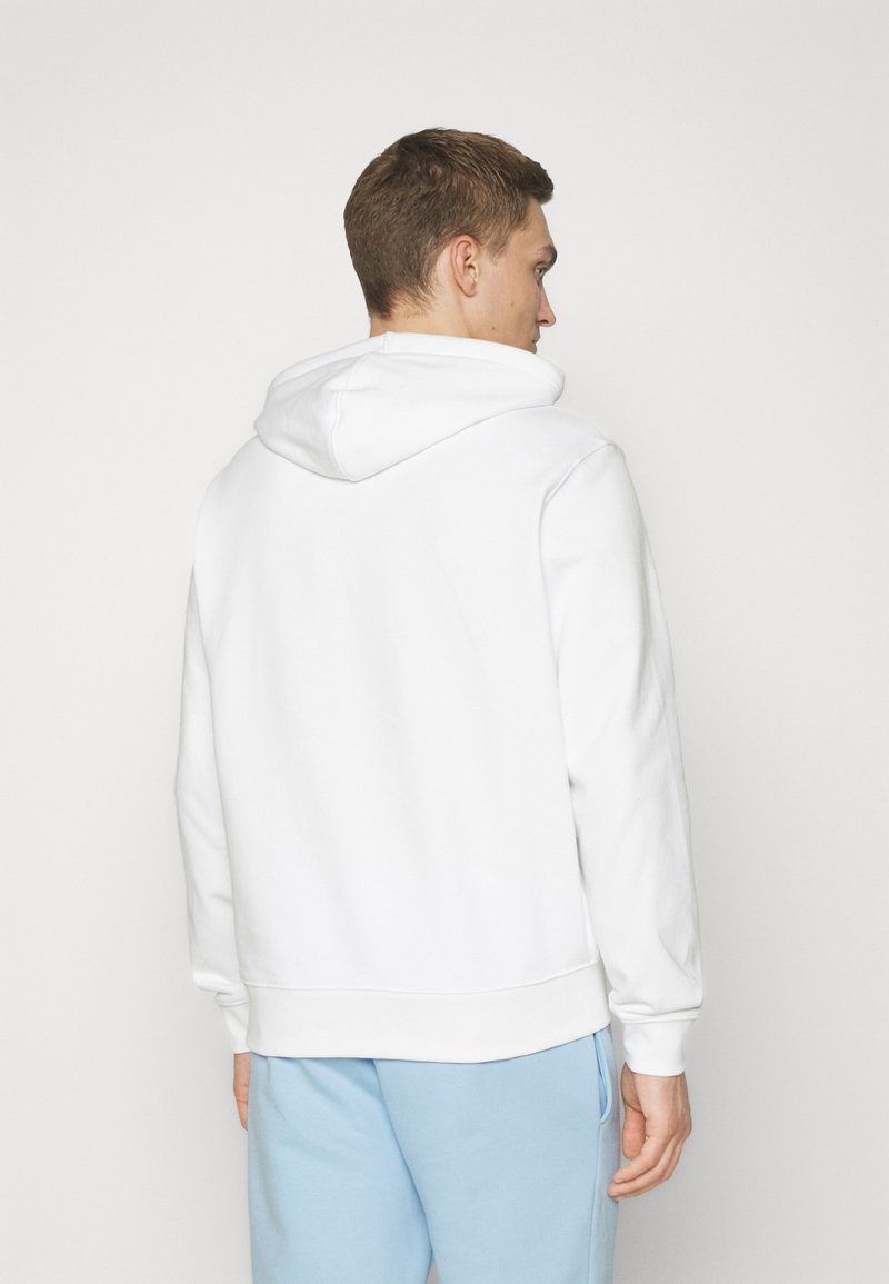 Sweatshirt à capuche blanche avec une coupe décontractée, des poignets et un ourlet côtelés. Texture lisse et sans motifs visibles. Porté avec un pantalon de jogging bleu clair.