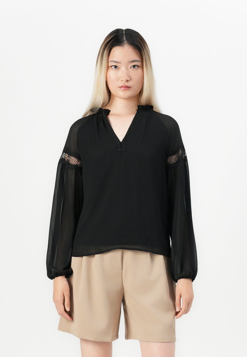 Donna con lunghi capelli ombre che indossa una camicetta nera trasparente con dettagli in pizzo e pantaloni corti beige fino al ginocchio, in piedi su uno sfondo bianco.