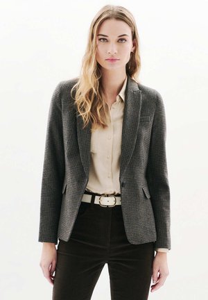 Veste en pied-de-poule grise à deux boutons, revers crantés et poches avant, associée à une chemise beige et un pantalon en velours côtelé marron.