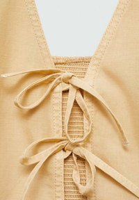 Beige Oberteil mit tiefem V-Ausschnitt, gerafftem Smocking-Detail und zwei Bindebändern vorne. Weicher, strukturierter Stoff mit Schnürungselementen.