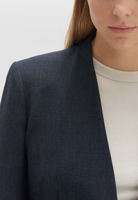 Blazer bleu marine en tissu texturé, avec un col cranté et des épaules structurées, associé à un haut blanc côtelé en dessous.