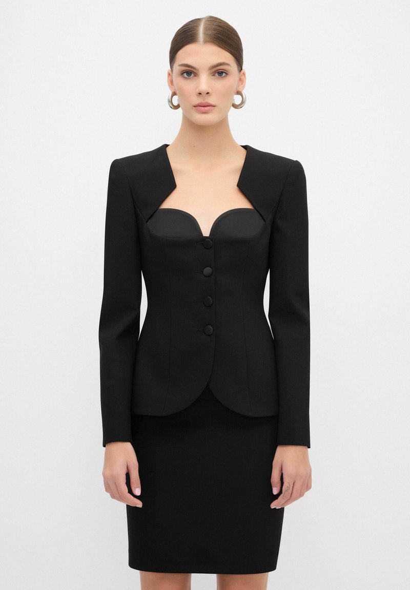 Blazer nero strutturato con corpino aderente, scollatura quadrata e tre bottoni, abbinato a una gonna a tubino. Tessuto liscio e spalle definite.