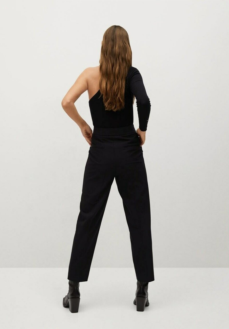 Femme aux longs cheveux bruns, debout les mains sur les hanches, portant un top noir à une épaule, un pantalon noir et des bottines noires à talons, vue de dos.