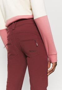 Ciżmowe spodnie Roxy w kolorze bordowym z dwoma zasuwanymi tylnymi kieszeniami, gładką strukturą, średnim stanem i dopasowanym krojem. Akcenty w kolorze różowym na górze.