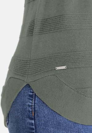 Nahaufnahme des Saums eines grünen Strickpullovers mit horizontaler Rippung und einem kleinen Metall-Brandtag über der Tasche einer blauen Jeans.