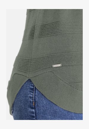 Nahaufnahme des Saums eines grünen Strickpullovers mit horizontaler Rippung und einem kleinen Metall-Brandtag über der Tasche einer blauen Jeans.